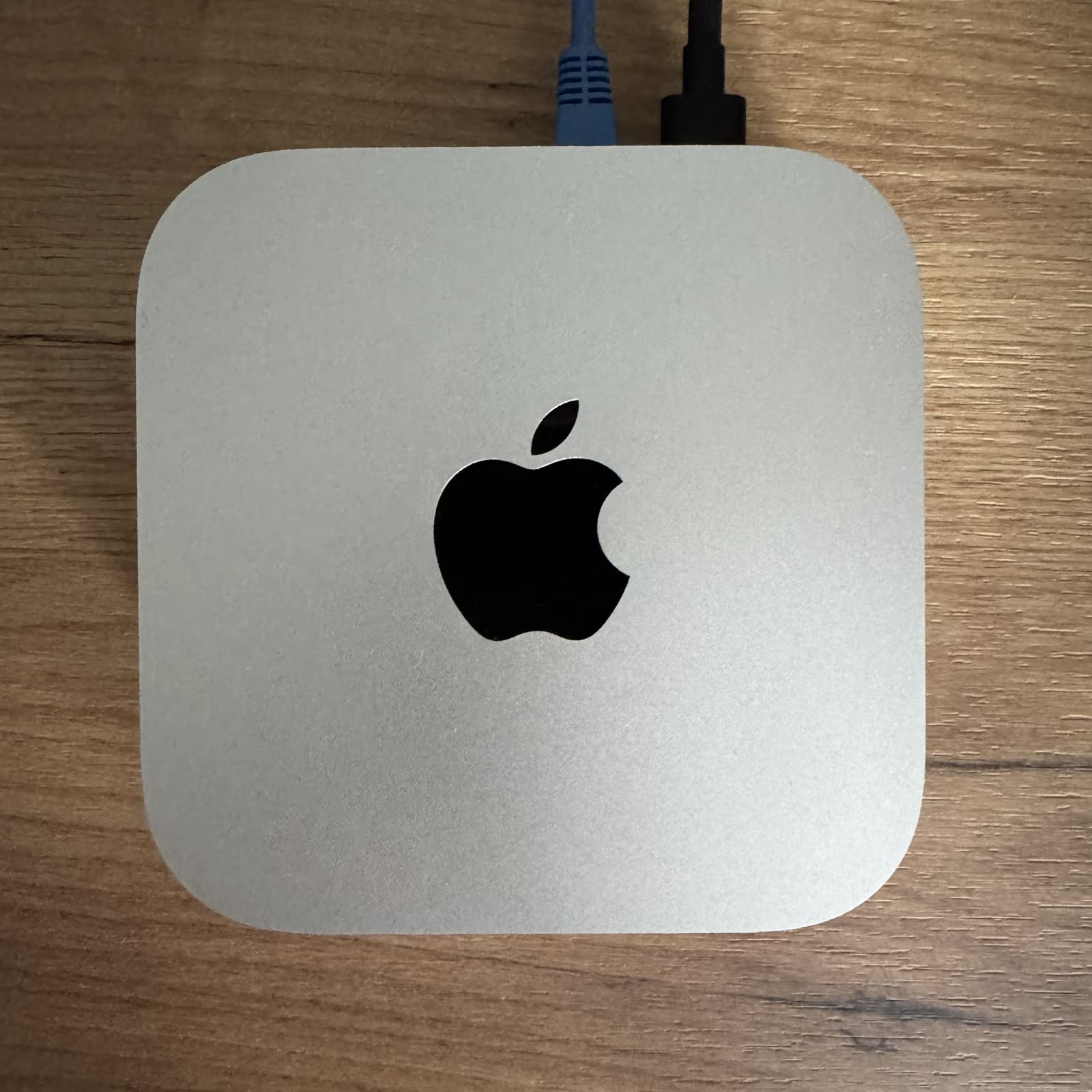 A silver Mac mini seen from above on a wooden desk, with a blue network cable and a black power cable plugged in at the back.