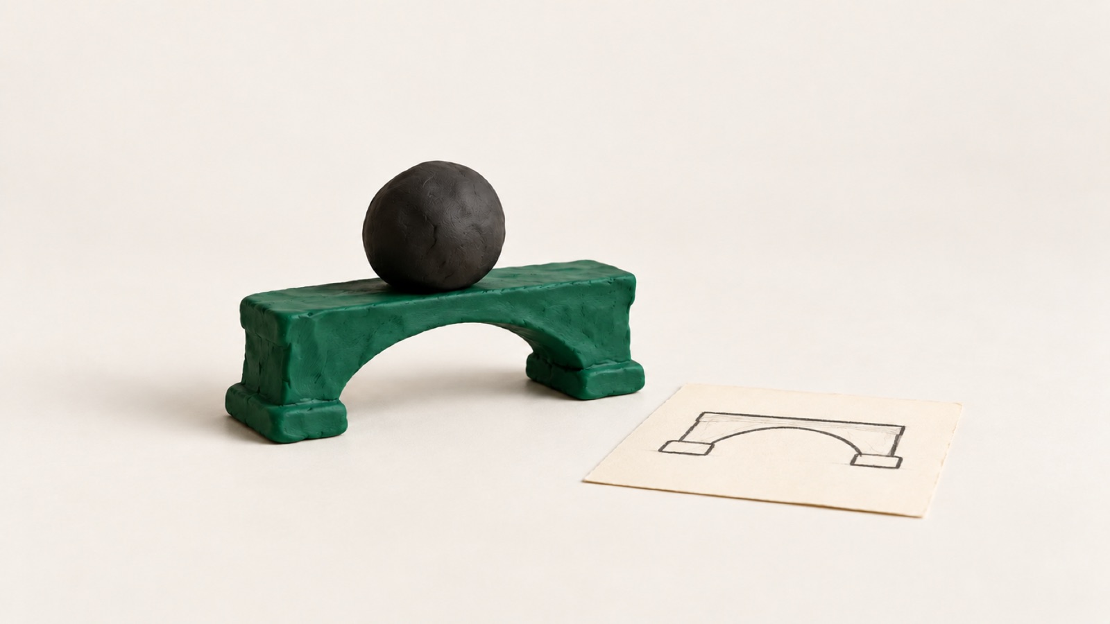 A small green clay bridge holds a heavy black clay ball. Next to it lies the paper sketch it was built from.